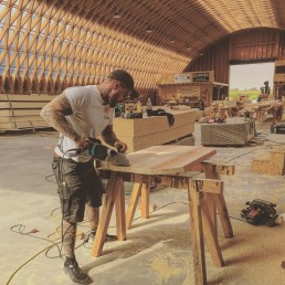 Ein Mann arbeitet an einem Holztisch in einer Werkstatt, umgeben von verschiedenen Werkzeugen und Geräten.